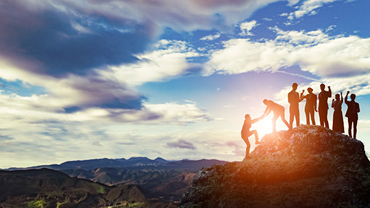 A group of people helping each other reach the top of a mountain at sunset, symbolizing collaboration and support.